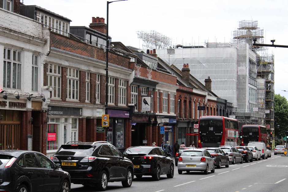 A street scene showing a row of brick and white-painted terraced houses with large windows, some with decorative molding, and a building under scaffolding on the right side, indicating ongoing construction or refurbishment. In front of the houses, a line of vehicles including several black, grey, and white cars are parked along the pavement. Two red double-decker buses, numbered 343 and 42, are visible on the street, with some pedestrians walking on the sidewalk. The pavement has a narrow curb, and there are street signs and traffic signals visible, including a 20 mph speed limit sign. The sky is overcast, and the scene captures typical urban activity associated with home relocation or furniture transport in a city environment, reflecting the logistics involved in house removals in an area like Lordship Lane, Dulwich, as managed by Dulwich Movers.