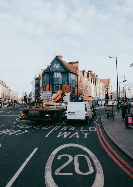 A white panel van and a flatbed truck are parked on a busy street in Dulwich, with the flatbed truck carrying wooden pallets and other packing materials. The vehicles are positioned near a pedestrian crossing with road markings indicating a 20 mph speed limit. In the background, there is a multi-story building with a dark blue facade and large windows, alongside other mixed-use buildings with brick and light-colored facades. Pedestrians are walking along the pavement, some carrying bags, and bicycles are parked to the side. Street lamps line the street, and the sky is partly cloudy. The scene captures the typical environment of a home relocation or furniture transport process, with Dulwich Movers possibly involved in the logistics of packing and moving households in the Lordship Lane area, illustrating the preparation and transport phase of a professional removals service.