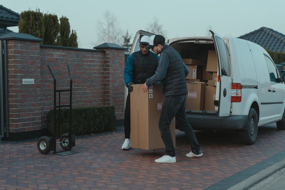 Two men are engaged in a house removal task, standing outside a residential property on a paved driveway. They are lifting and positioning a large cardboard box, with one wearing a black quilted vest, grey hoodie, and white sneakers, and the other in a blue and black jacket with gloves. Behind them, a white van with its rear doors open is loaded with several packed cardboard boxes, some sealed with packing tape, and visible within the vehicle are additional boxes and packaging materials. A black utility trolley is positioned nearby, ready for transporting items, and the scene is set in a suburban area with a brick wall, hedges, and residential rooftops in the background. The lighting suggests a cloudy day, emphasizing the practical nature of furniture transport and packing during a home relocation process. This scenario illustrates the logistics involved in removals, with Dulwich Movers providing professional assistance with packing and moving services in East Dulwich SE22.