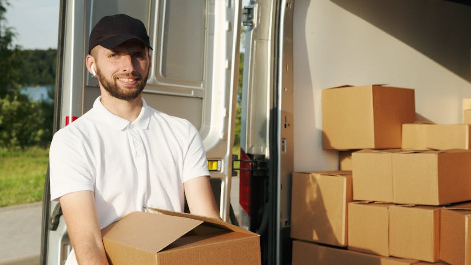 A man with a beard and wearing a black cap and white polo shirt is standing outside of a moving van during a home relocation process. He is holding a cardboard box while standing near the open side of the van, which is filled with several other packed cardboard boxes stacked neatly inside. The van is parked on a paved area, with a green landscape and trees visible in the background, under clear daylight. Moving blankets, straps, or other loading equipment are not visible in this image. The scene depicts typical furniture transport and packing activities associated with professional removals by Dulwich Movers, aligned with house removals and moving logistics in East Dulwich SE22.