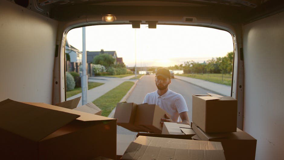 Two men are engaged in a house removal task, standing outside a residential property on a paved driveway. They are lifting and positioning a large cardboard box, with one wearing a black quilted vest, grey hoodie, and white sneakers, and the other in a blue and black jacket with gloves. Behind them, a white van with its rear doors open is loaded with several packed cardboard boxes, some sealed with packing tape, and visible within the vehicle are additional boxes and packaging materials. A black utility trolley is positioned nearby, ready for transporting items, and the scene is set in a suburban area with a brick wall, hedges, and residential rooftops in the background. The lighting suggests a cloudy day, emphasizing the practical nature of furniture transport and packing during a home relocation process. This scenario illustrates the logistics involved in removals, with Dulwich Movers providing professional assistance with packing and moving services in East Dulwich SE22.