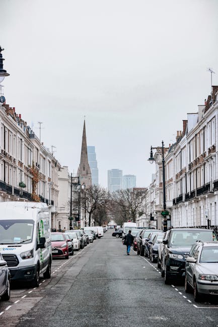 A view of a residential street during daytime showing parked cars lining both sides of the road, with a van positioned on the left side, potentially for home relocation purposes. In the background, th