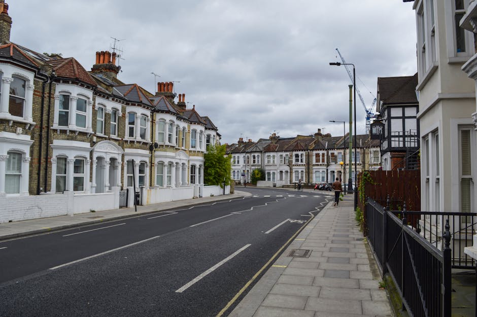 A view of a residential street during daytime showing parked cars lining both sides of the road, with a van positioned on the left side, potentially for home relocation purposes. In the background, th