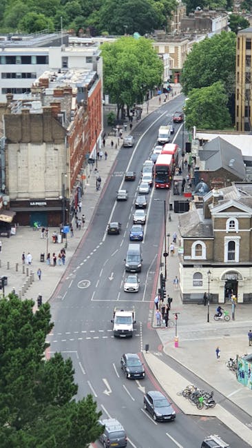 An aerial view of a busy street in West Dulwich SE21 with a mix of vehicles including cars, a red double-decker bus, and bicycles. The street is lined with trees providing greenery, and there are pedestrians walking along the sidewalks. Several multi-storey buildings with brick and modern facades are visible, along with a small church-style building at the corner. The scene captures the typical urban environment during daylight hours, with the roads showing signs of ongoing traffic and movement. This image relates to house removals and relocation services offered by Dulwich Movers, highlighting the logistics involved in furniture transport and home relocation through urban streetscape and traffic management.
