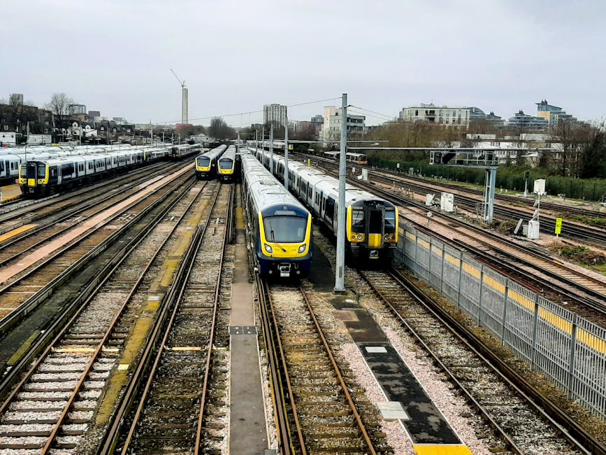 A view of multiple modern commuter trains with yellow and blue livery on railway tracks at a station in West Dulwich SE21. The image shows several train sets parked side by side on parallel tracks, with some trains heading towards the distance and others positioned closer to the platform area. The tracks are made of metal rails mounted on wooden sleepers, with gravel ballast surrounding them. A concrete platform is visible to the left, possibly where passengers wait for services, though no people are present. Overhead electrical wires run above the tracks, supported by metal poles. In the background, there are several mid-rise buildings, some with balconies, along with leafless trees and a cloudy sky indicating overcast weather. The scene demonstrates the railway environment associated with local transportation services, which often coordinate with house removals or relocation activities by Dulwich Movers to assist clients in moving belongings during house relocations near North Dulwich station.